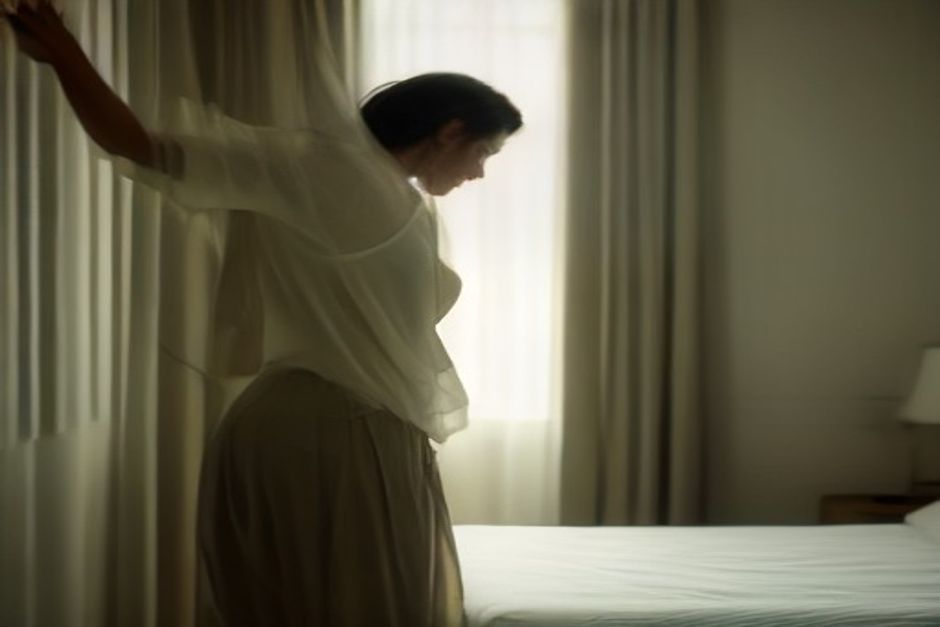A person stretching in a standing forward bend beside a hotel bed, soft morning light through curtains, earth-toned room