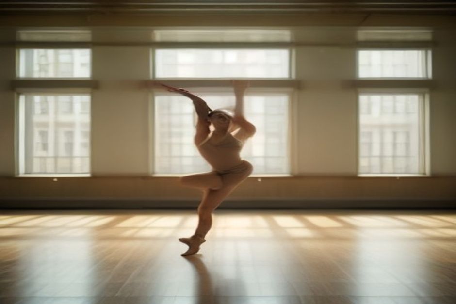 A dancer seated in a wide-legged forward fold on a wooden studio floor, warm natural light through tall windows