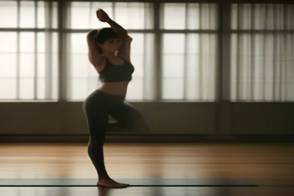 A person folding forward in a standing pose on a mat in a warm, humid studio with soft light